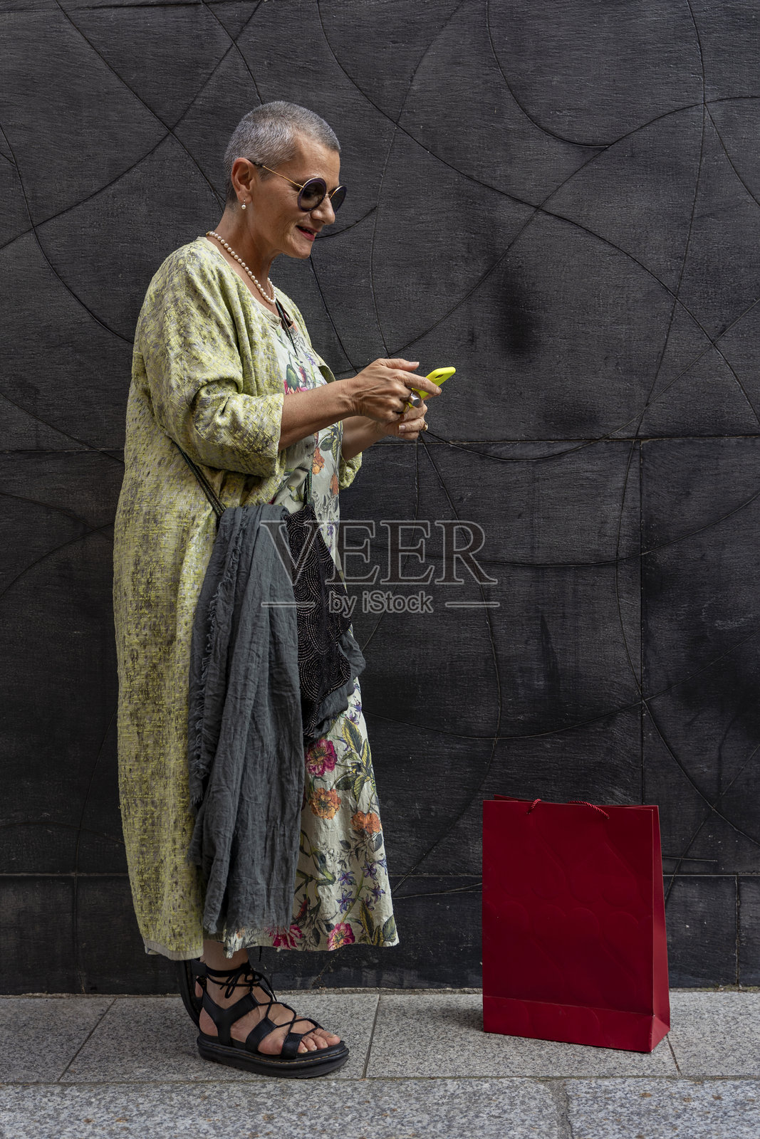 优雅微笑的成熟的 女性，剃光灰色头发，站在黑色墙前用手机发短信。照片摄影图片