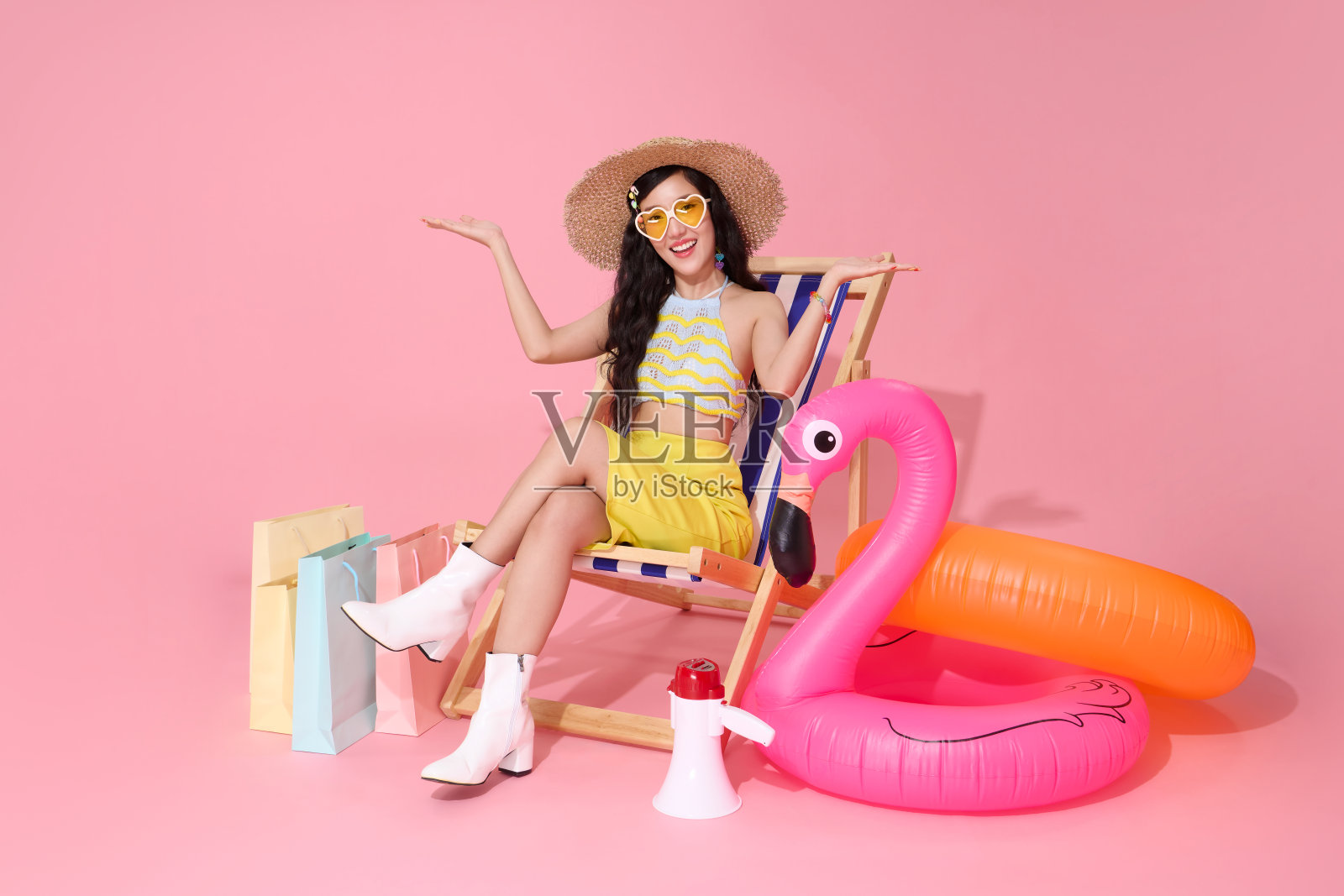 戴草帽和心形太阳镜的女人坐在沙滩椅上，双臂伸展，手里拿着购物袋，背景为粉色。非常适合夏季促销。照片摄影图片