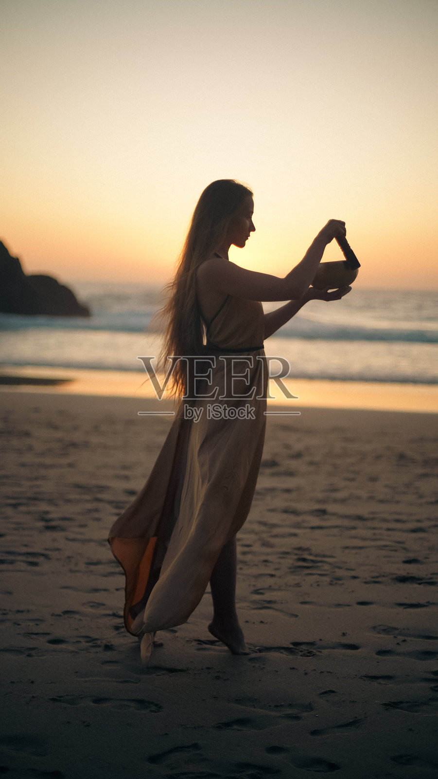 穿着裙子的女孩在日落时分的海洋背景下，手持音乐碗的剪影。水分补充。柔焦效果。照片摄影图片