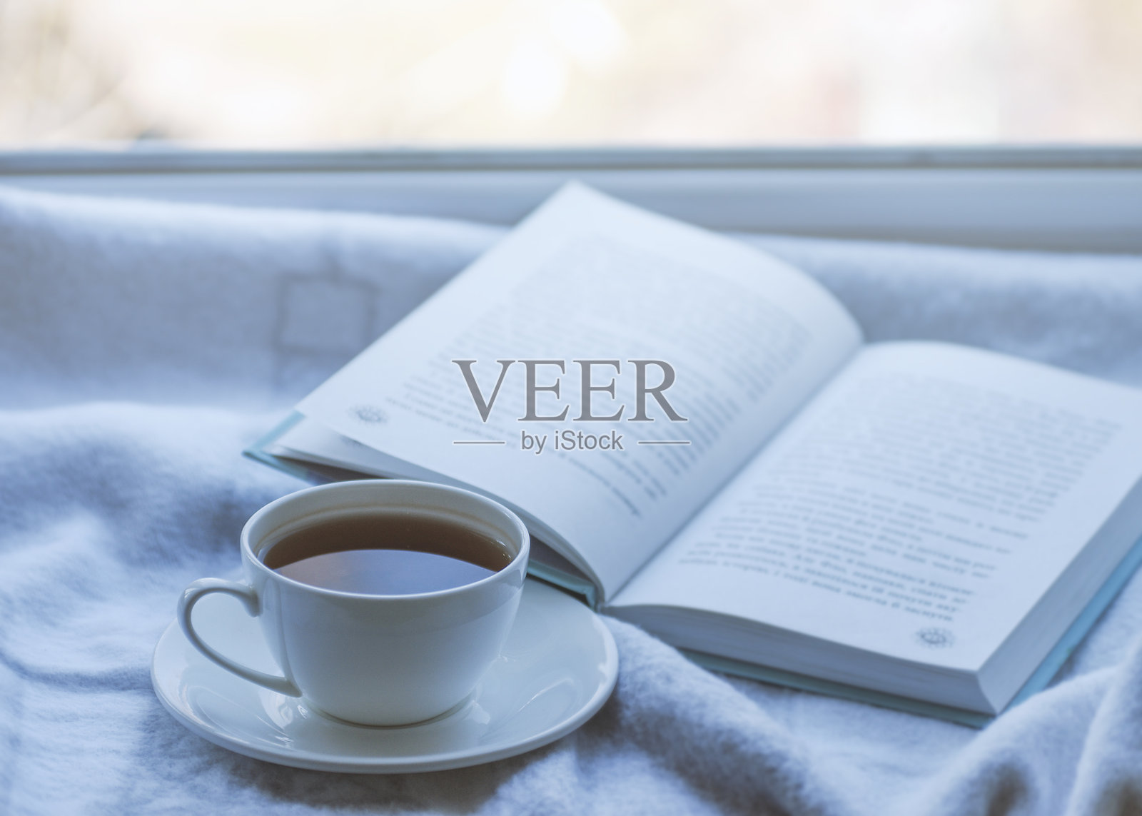 温馨的冬季静物：窗台上放着一杯热茶和一本书，旁边有温暖的格子毯，窗外是雪景。照片摄影图片