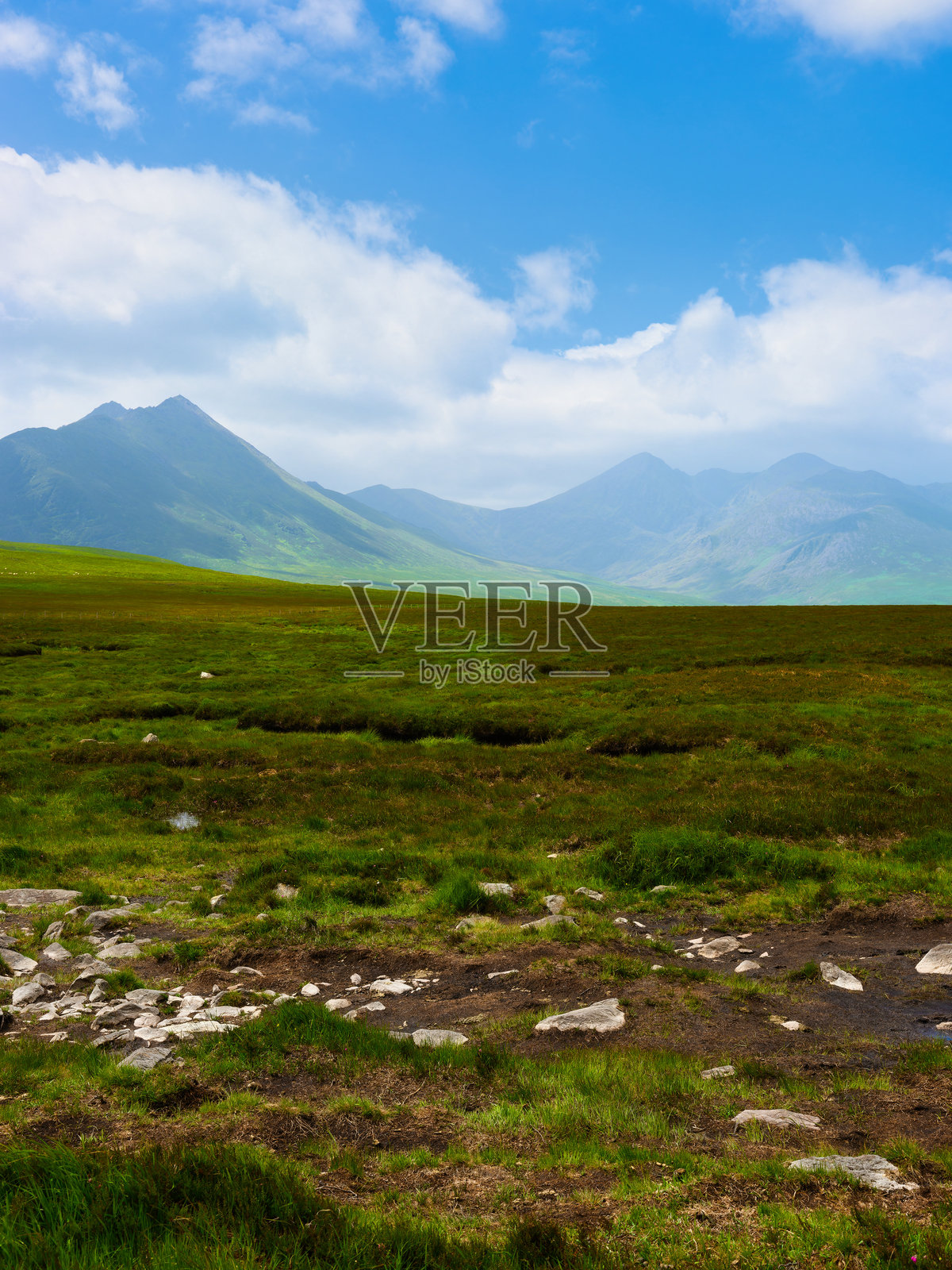 前景是起伏的绿色田野，延伸至爱尔兰壮丽的山脉。天空湛蓝，点缀着几朵白云。照片摄影图片