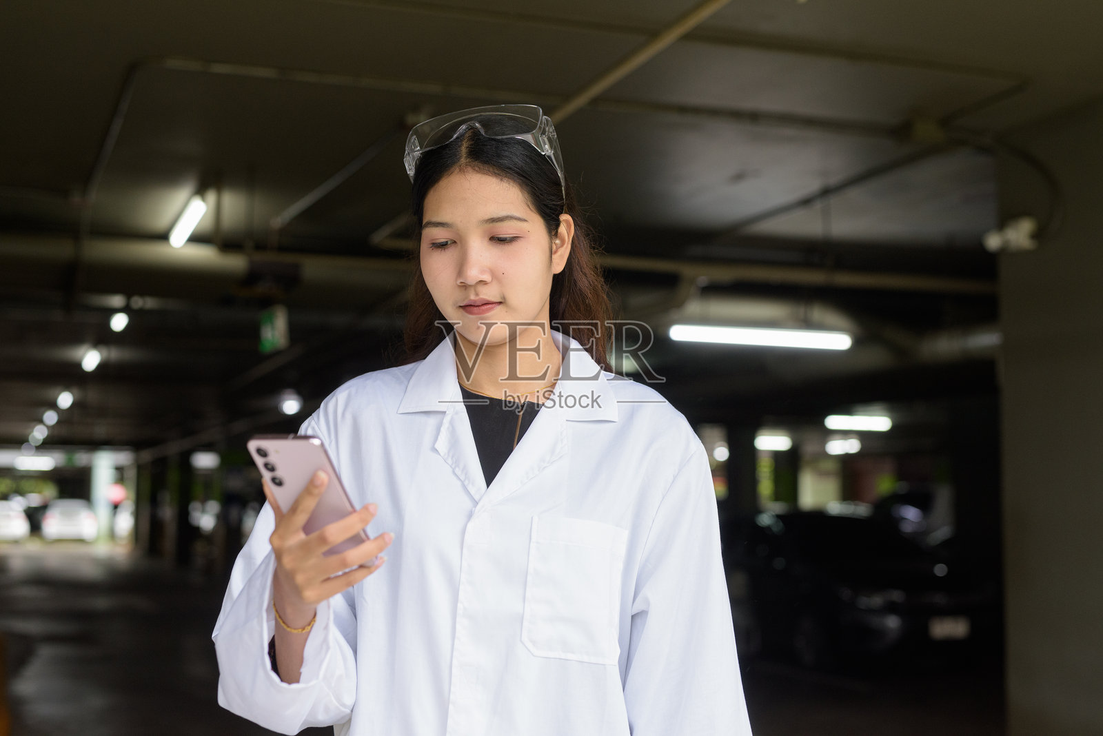 穿着实验室外套和护目镜的女科学家在建筑外用手机发短信照片摄影图片