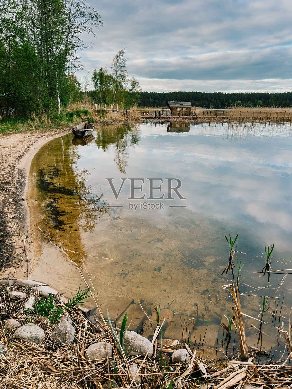 木船在平静的湖水中倒影，背景有码头和小木屋，周围被植被环绕，天空多云。乡村小屋坐落在浓密的绿树丛中，天空阴沉。照片摄影图片