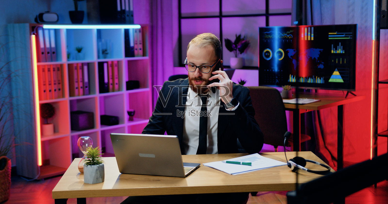 办公室工作流程中，一位英俊、忙碌、目标明确、技术娴熟的年轻男士，身穿正式服装，戴着眼镜，一边打电话，一边查看纸质报告和电脑信息。照片摄影图片