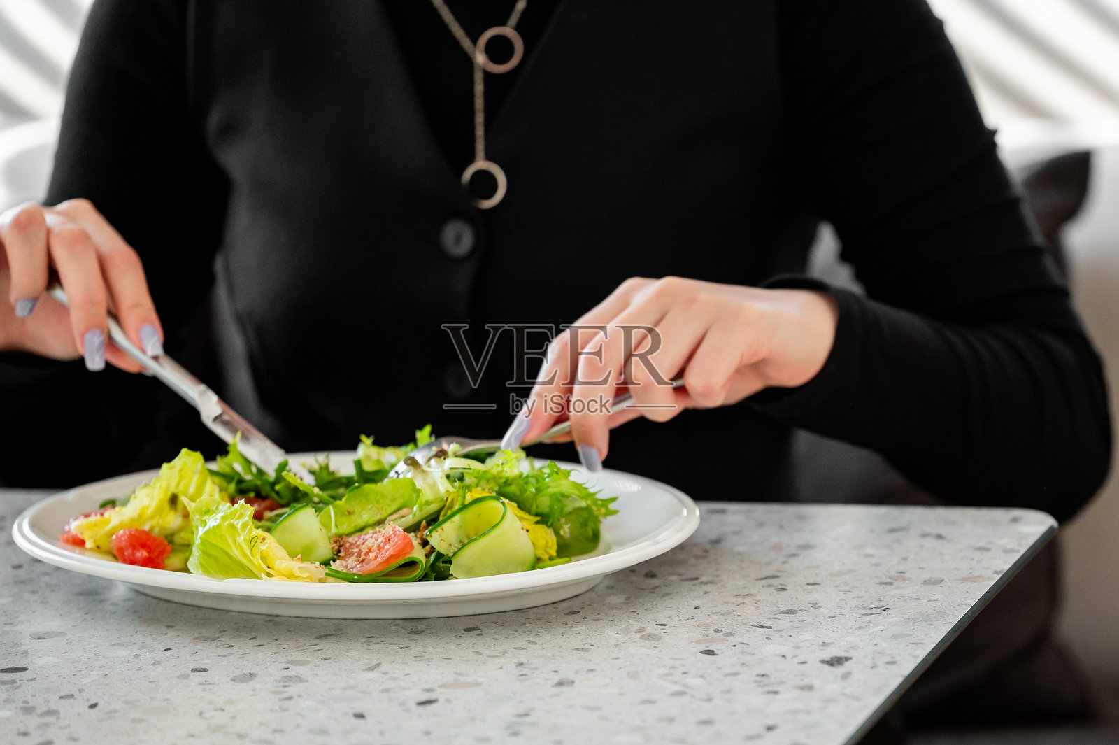 穿着黑色服装的女性用刀叉吃新鲜的蔬菜沙拉，特写手部在现代厨房桌子上，宣传健康生活方式和均衡营养。照片摄影图片