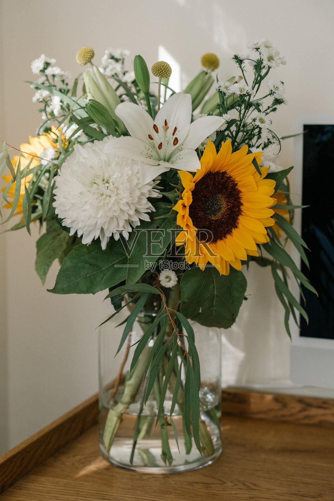 桌子上的玻璃花瓶里插着一束夏季花卉 - 百合、向日葵、花朵。夕阳的光线。照片摄影图片