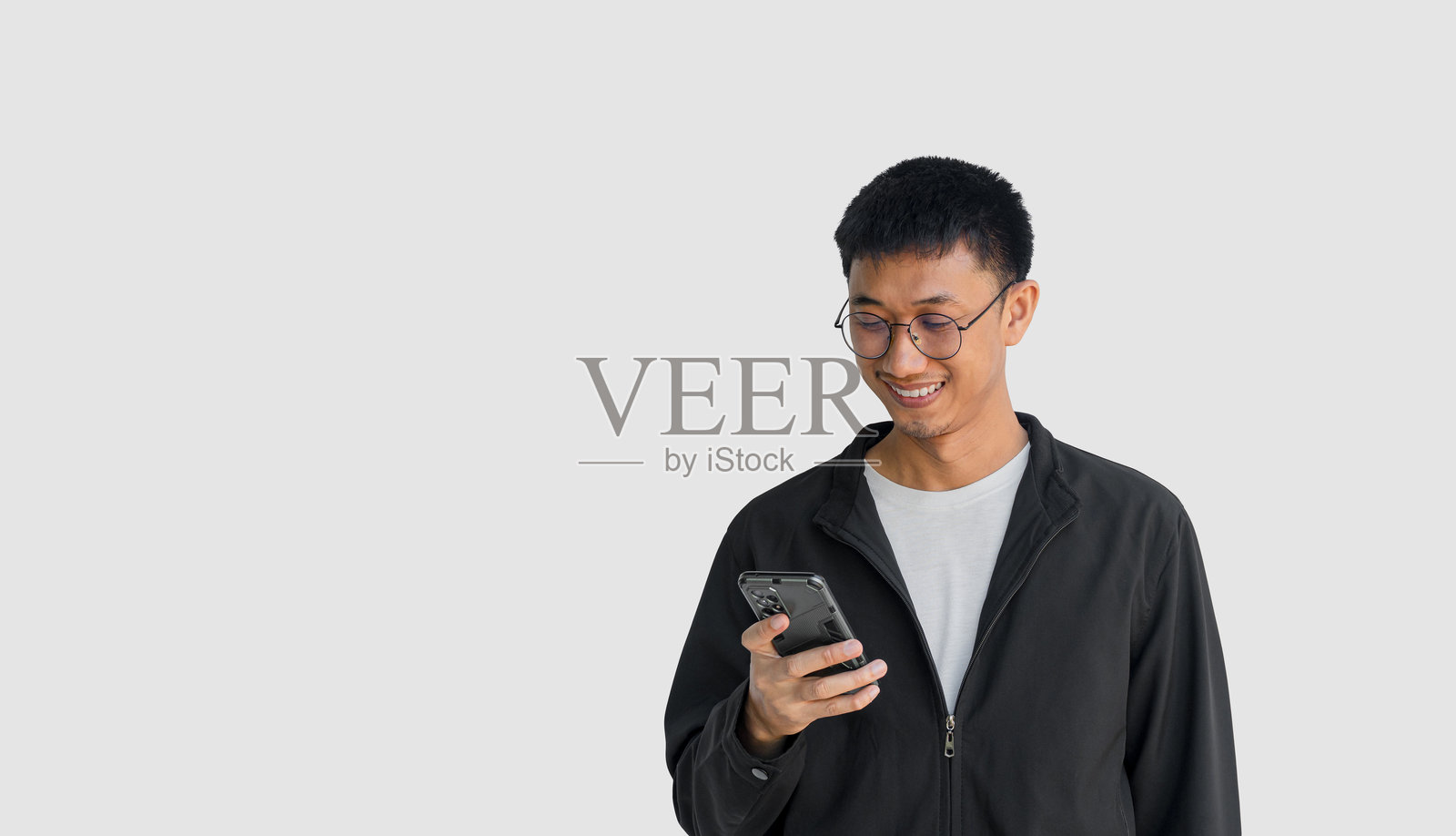 一位20多岁的年轻亚洲男子戴着眼镜，穿着黑色外套，愉快地在灰色背景下玩手机。通过智能手机与朋友聊天和打字，享受在线社交。照片摄影图片