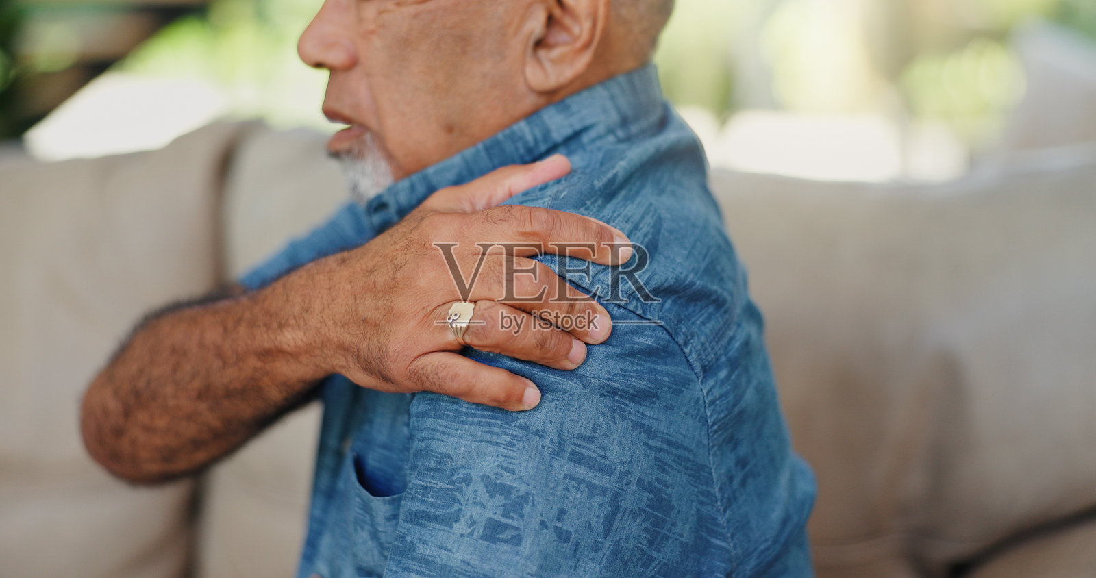 老年男性坐在沙发上，双手搭在肩膀上，感到肌肉紧张、关节炎或骨质疏松。老年人在家中因肌腱炎、神经损伤和扭伤而感到手臂受伤和关节疼痛。照片摄影图片