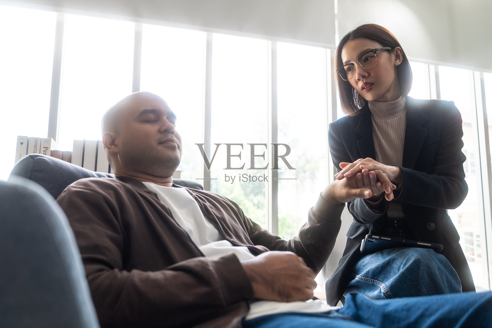 亚洲男子坐在沙发上，与亚洲女性心理医生在心理健康诊所进行咨询。心理医生正在交谈，并握住男性患者的手以支持他克服抑郁。照片摄影图片