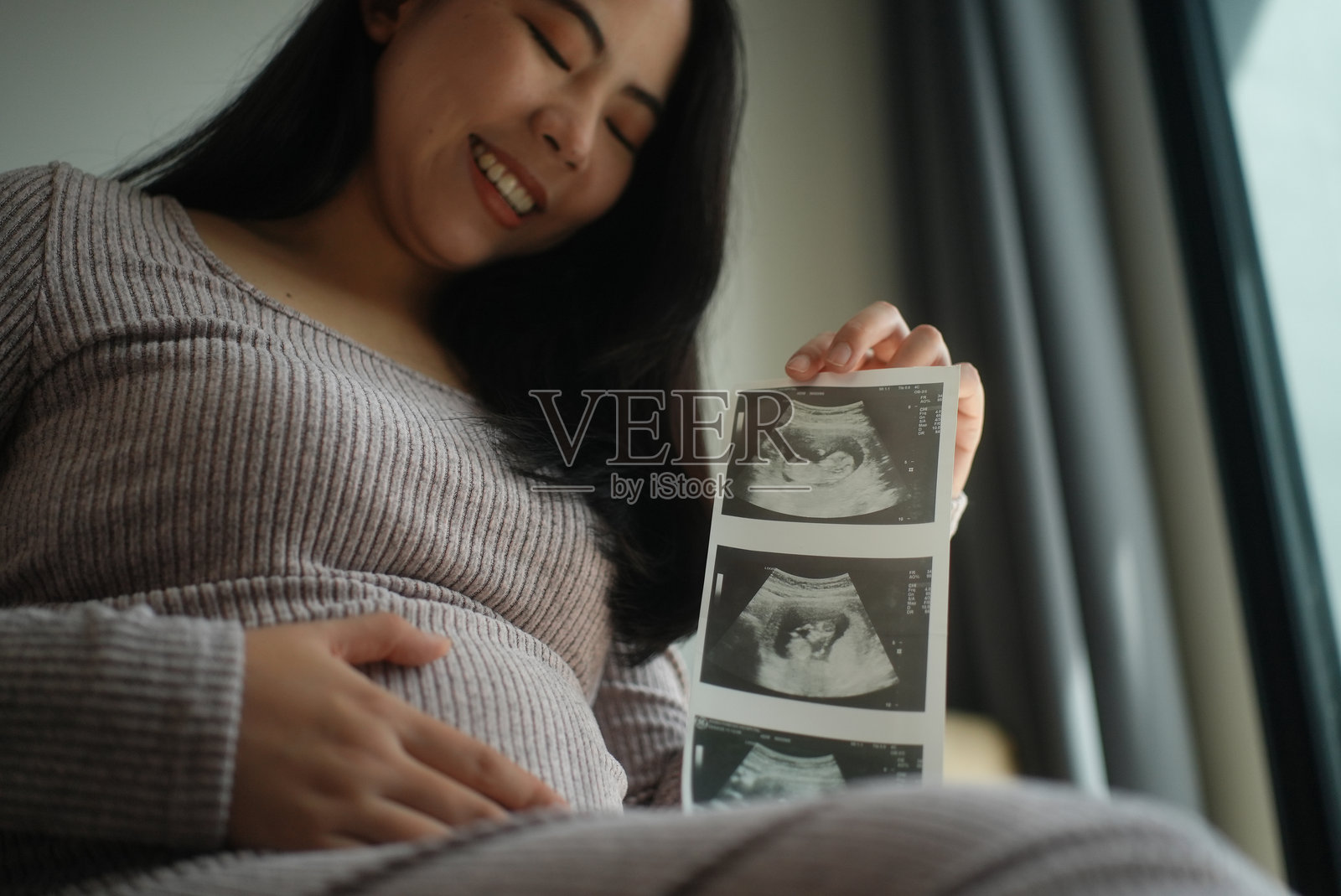 特写孕妇手持超声波扫描图，穿着休闲服坐在沙发上，幸福期待母亲身份，孕期准备迎接新生命的概念。照片摄影图片