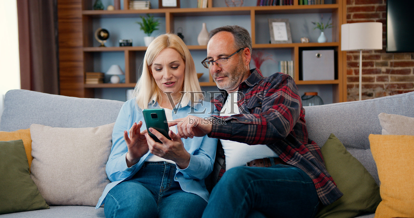 成熟夫妇在家中一起使用智能手机并微笑。男女在沙发上放松，网上购物，观看搞笑视频，使用社交媒体。照片摄影图片
