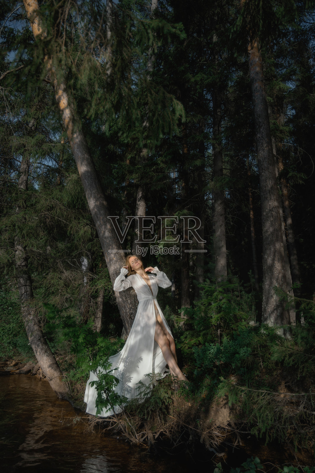 一位女性优雅地站在河边，身穿白色连衣裙，周围是高大的树木。阳光透过树叶洒下，营造出宁静的氛围。自然与时尚和谐交融。照片摄影图片