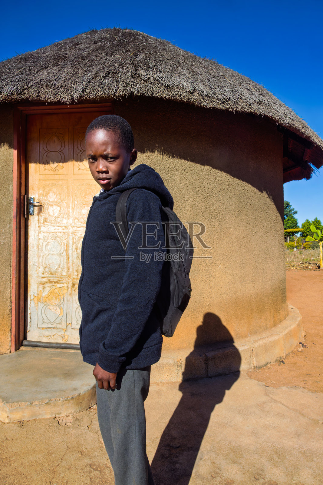 村庄里一个穿着校服的年轻非洲男孩站在房子前的院子里，房顶是茅草屋顶。照片摄影图片