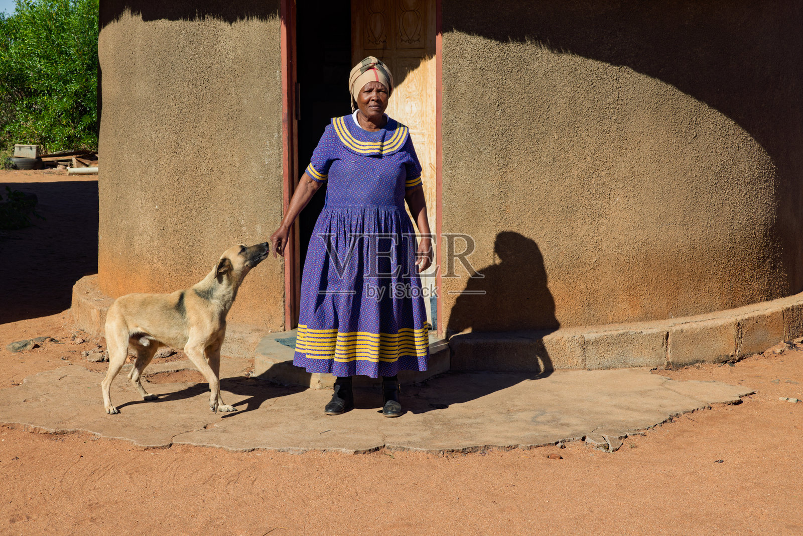 非洲村庄，单身老妇人戴着头巾，站在房子前面，狗在玩耍照片摄影图片