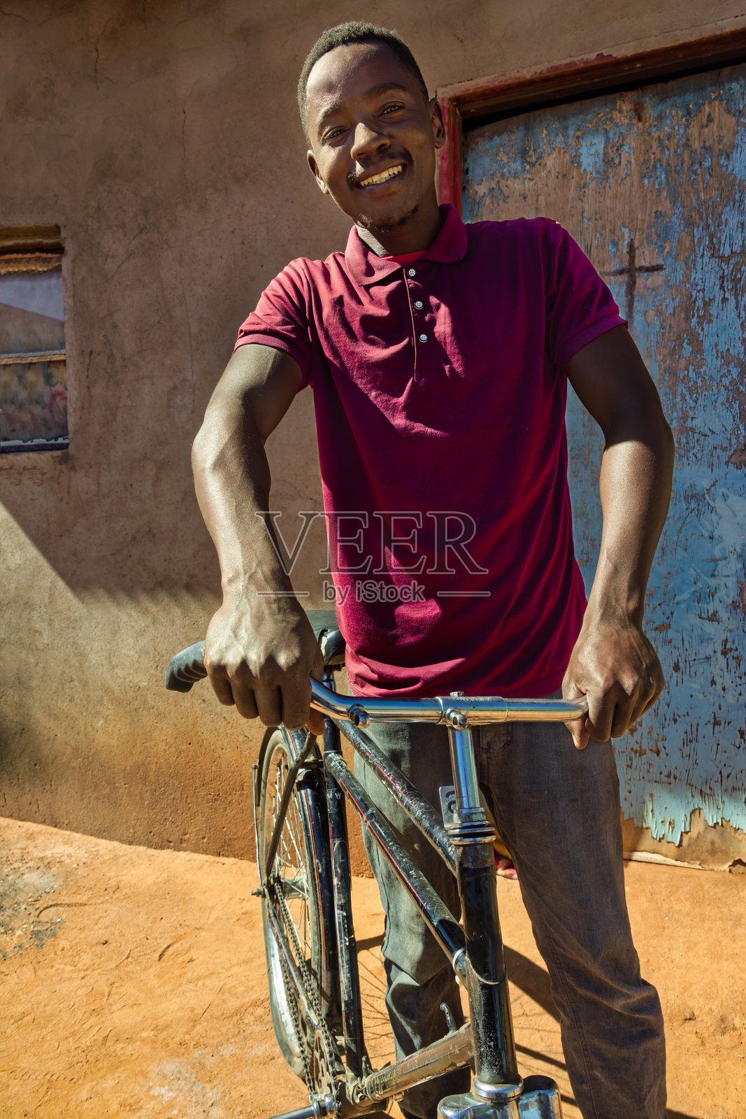 非洲村庄，一位年轻的非洲男子骑着自行车微笑，站在院子里的房子前，蓝色的门。照片摄影图片
