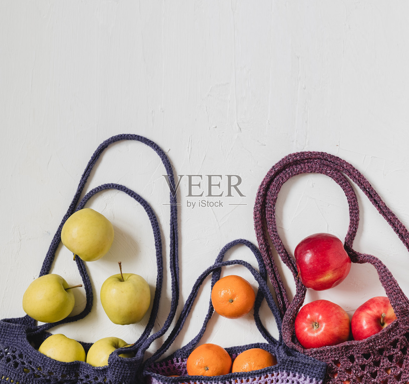 三个钩针环保袋，装有红苹果、金苹果和橘子。紫色和蓝色的网袋里装着有机苹果，背景为白色。环保购物。健康饮食理念。无塑料理念。零浪费。可持续生活方式。照片摄影图片