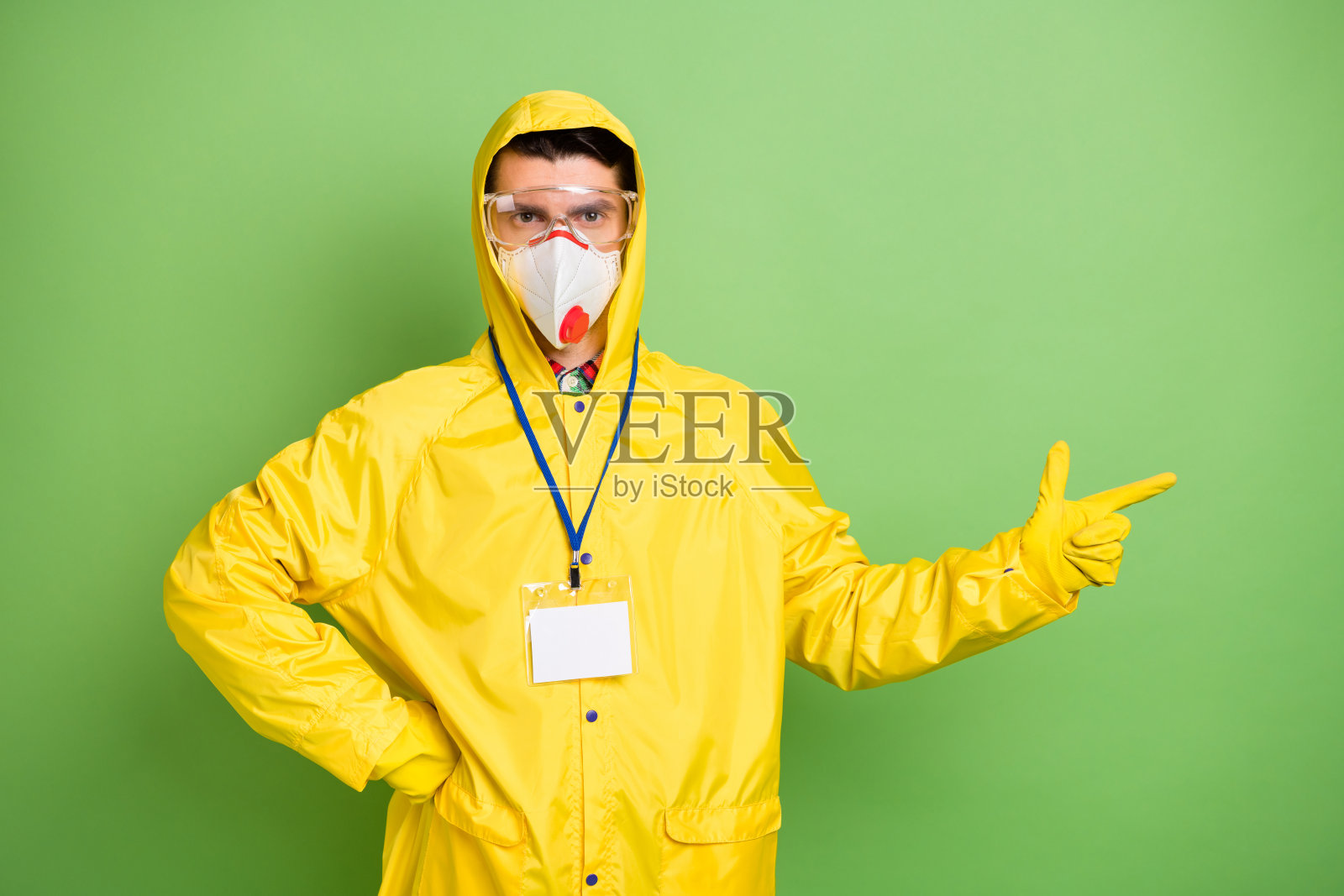 年轻男子戴着防护口罩、手套和护目镜，手指指向空白区域，建议隔离，背景为绿色。照片摄影图片