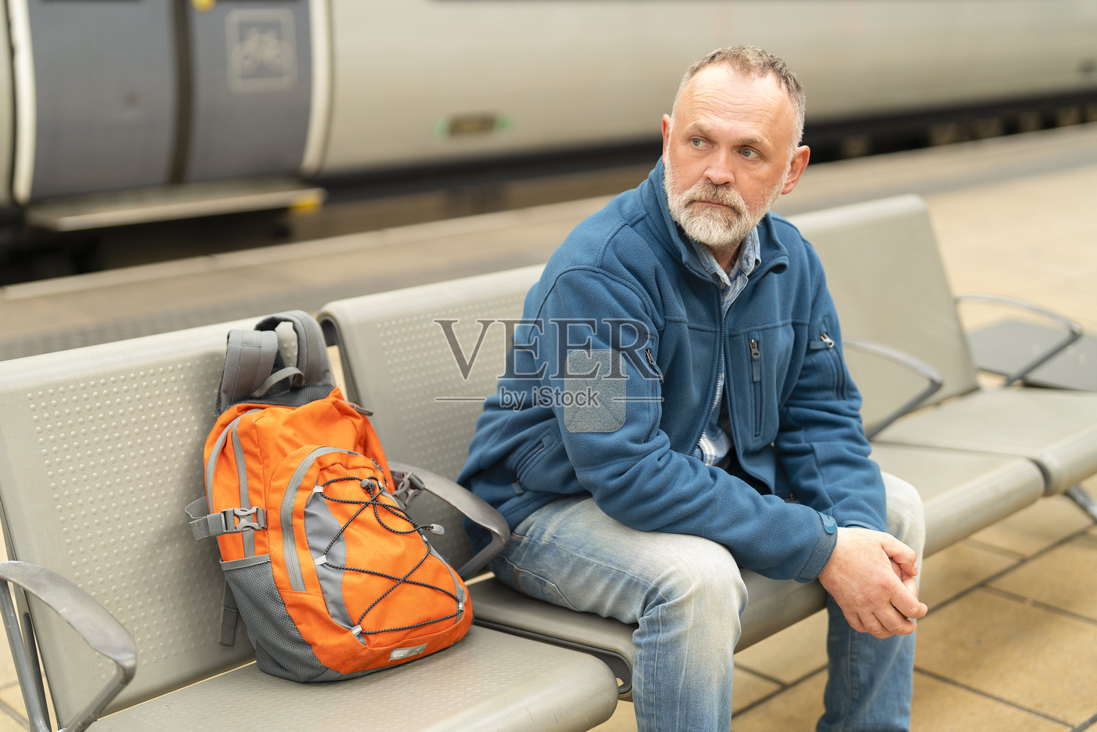 中年男子坐在城市火车站的长椅上，背着橙色背包，白天等待火车。照片摄影图片