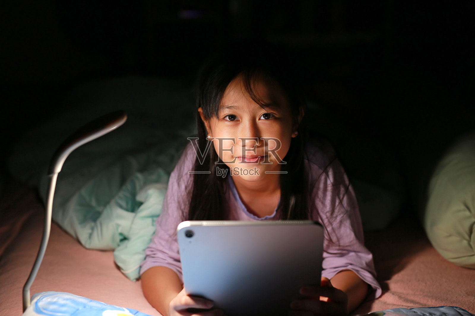 夜晚，亚洲女孩在床上躺着玩数字平板电脑的肖像。照片摄影图片