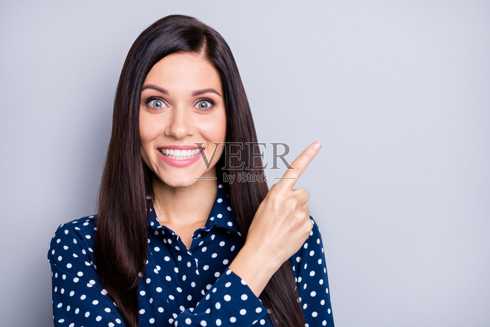 乐观的棕发女孩穿着蓝色上衣，指向空白处，背景为灰色。照片摄影图片