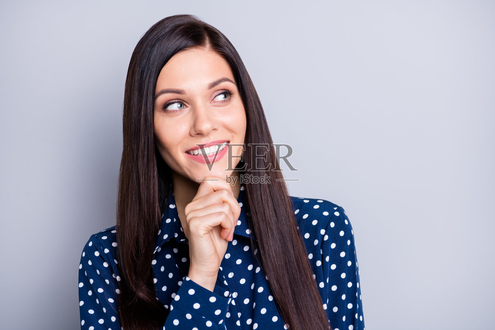 乐观的棕发女孩的肖像，手握下巴凝视空旷的空间，穿着蓝色上衣，背景为灰色。照片摄影图片