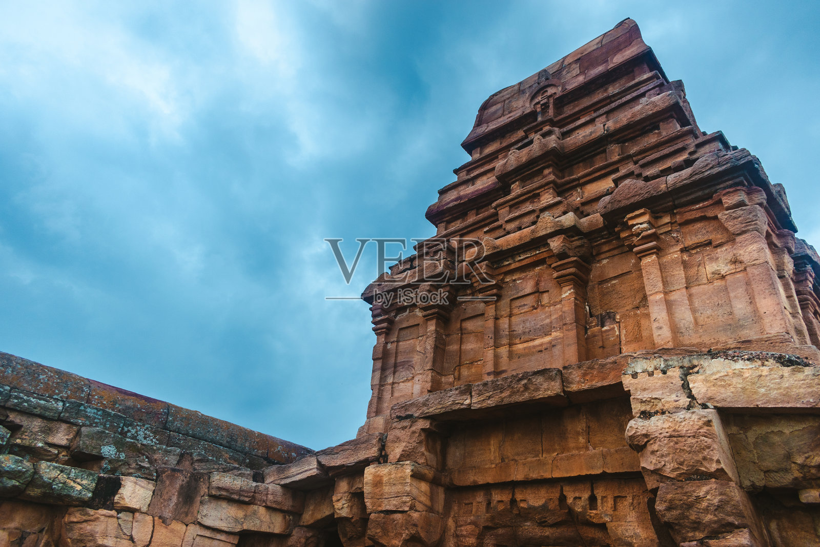 孤独的巴达米寺庙矗立在多变的天空下，周围是崎岖的地形。照片摄影图片