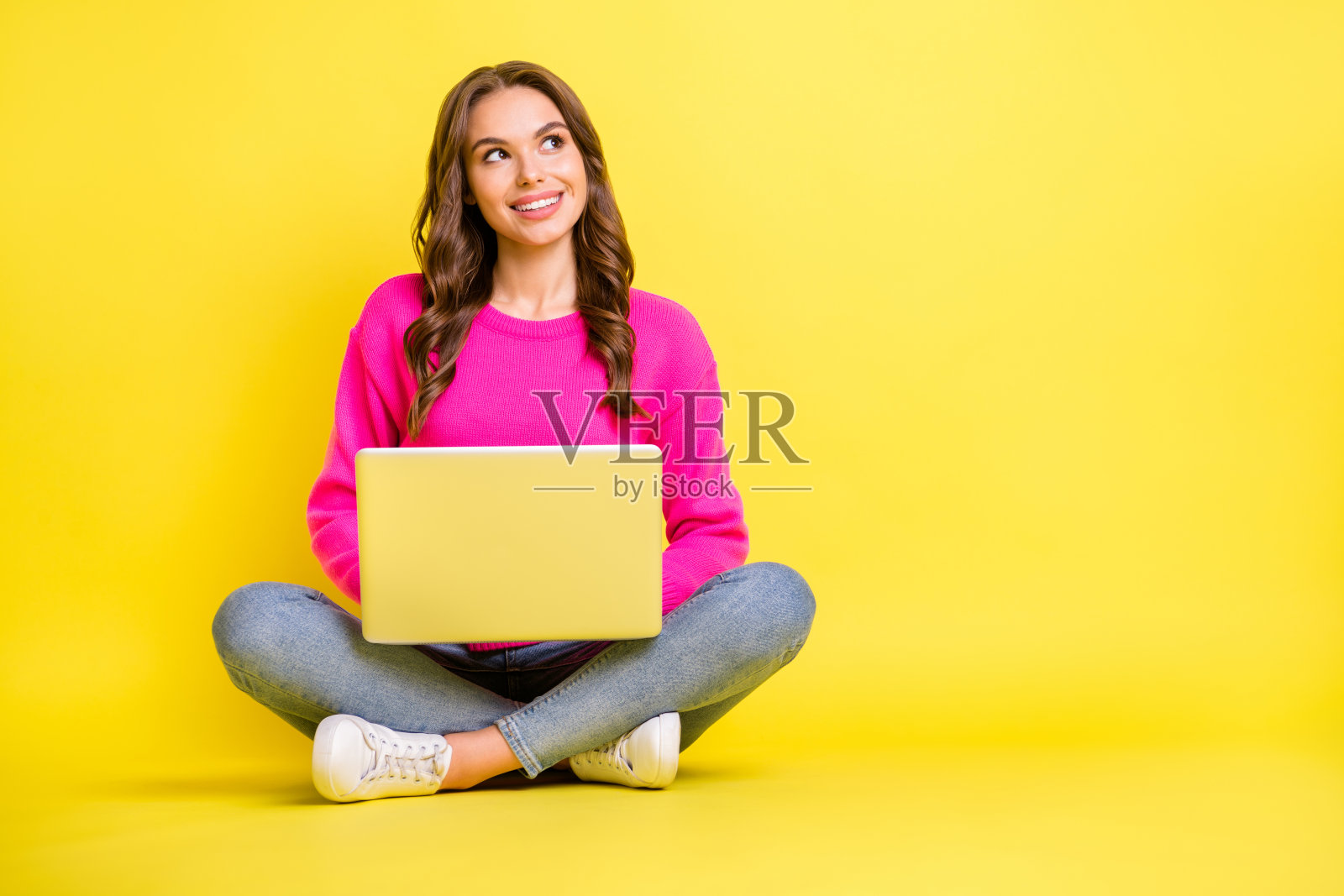 全身照，年轻快乐积极的微笑女孩坐在地板上，望着空白处，使用笔记本电脑，背景为黄色。照片摄影图片