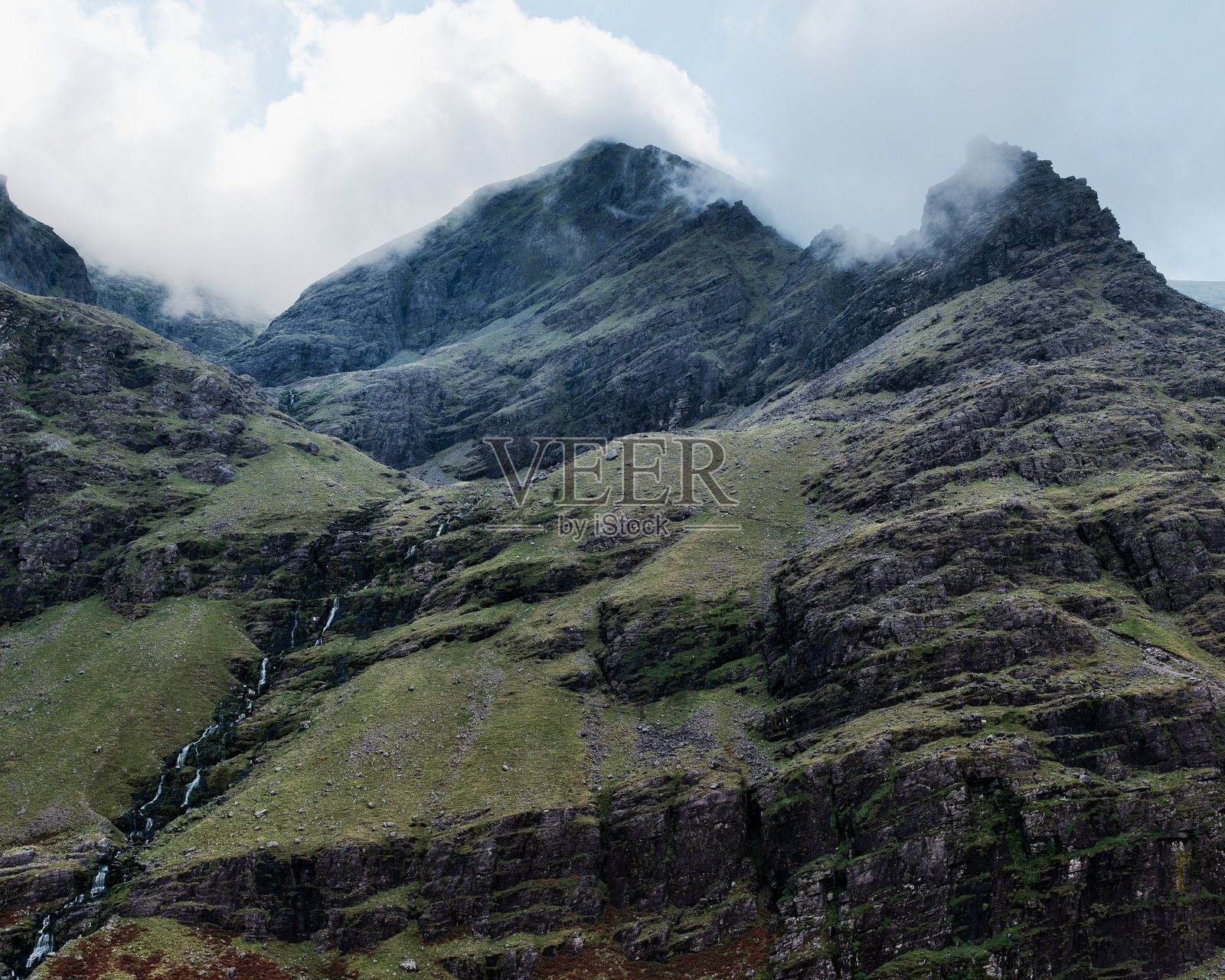 陡峭的岩石山坡展示了风化的石头。几条小瀑布涓涓而下。高处有低矮的云层。照片摄影图片