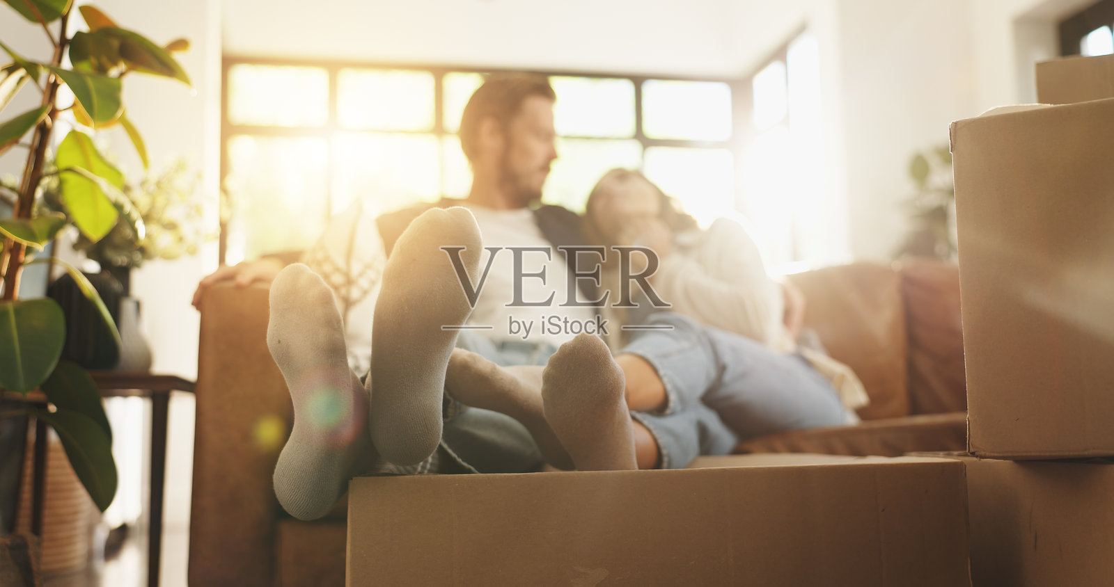 情侣在新家放松，脚边有搬家箱子，象征着搬迁、投资和共同的亲密关系。人们、镜头光晕和沙发代表着搬迁财产、贷款成功和对拥有关系的承诺。照片摄影图片