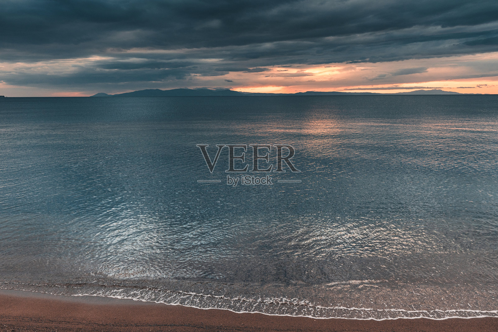 平静的海浪轻轻拍打着沙滩，背景是戏剧性的日落、乌云和岛屿。照片摄影图片
