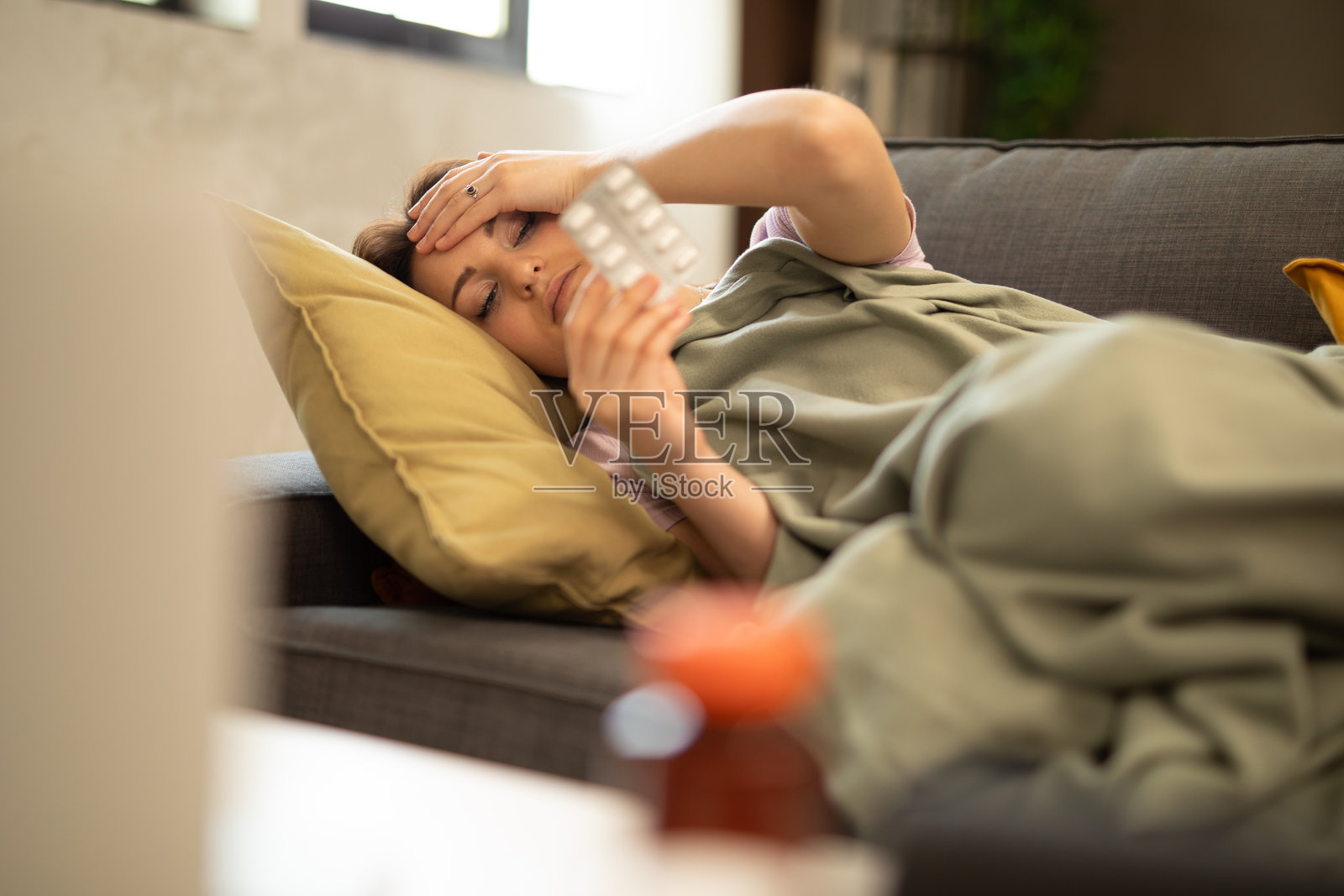 生病的女人躺在沙发上，手里拿着药片，额头贴着手。照片摄影图片