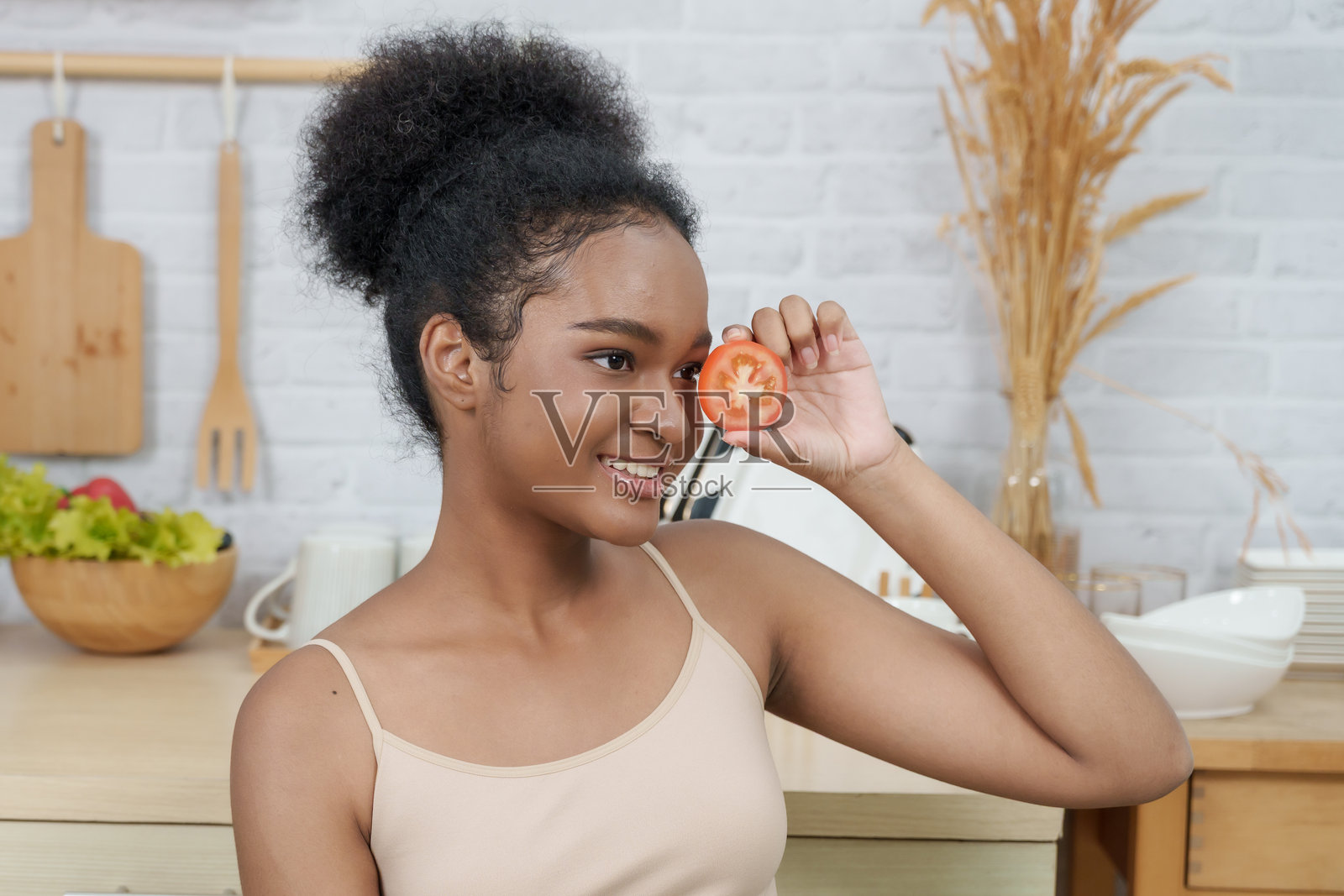 非洲少女在厨房里微笑着将半个番茄靠近脸庞，宣传自然美、护肤和通过清洁饮食与蔬菜滋养肌肤的健康生活方式。照片摄影图片