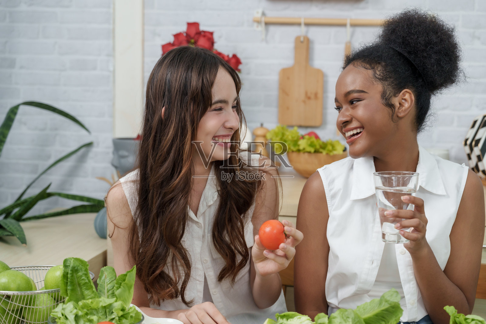 多种族的少女们在厨房享受护肤生活方式，健康饮食，非洲女孩手持水杯和番茄，亚洲朋友微笑，倡导自然美和肌肤健康。照片摄影图片