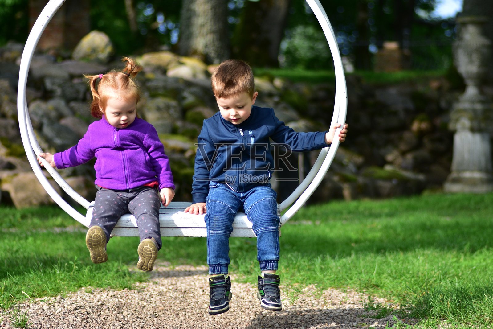 两个小孩在阳光明媚的公园里一起坐在白色金属秋千上。快乐的兄弟姐妹或朋友们穿着色彩鲜艳的外套，享受着愉快的玩耍时光。明亮而快乐的童年时刻。照片摄影图片
