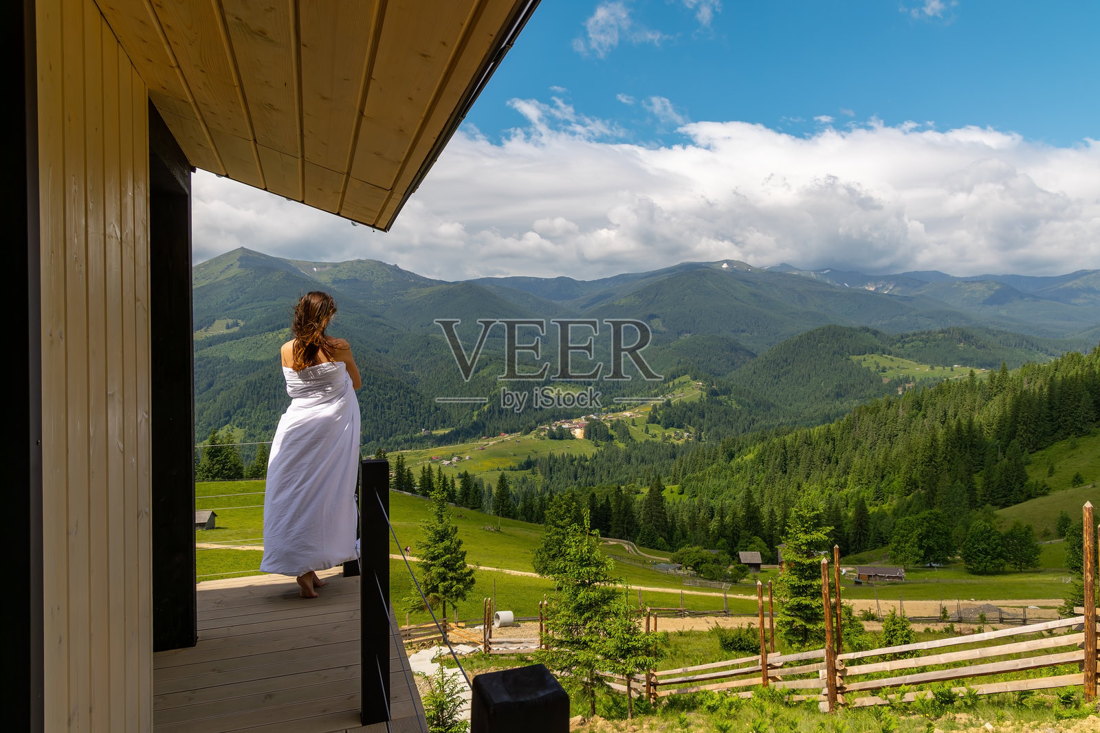 裹着白色毛毯的女人俯瞰绿色山谷。一个赤脚的女人裹着白色毛毯，站在木屋的阳台上，凝视着广阔的绿色山谷。照片摄影图片