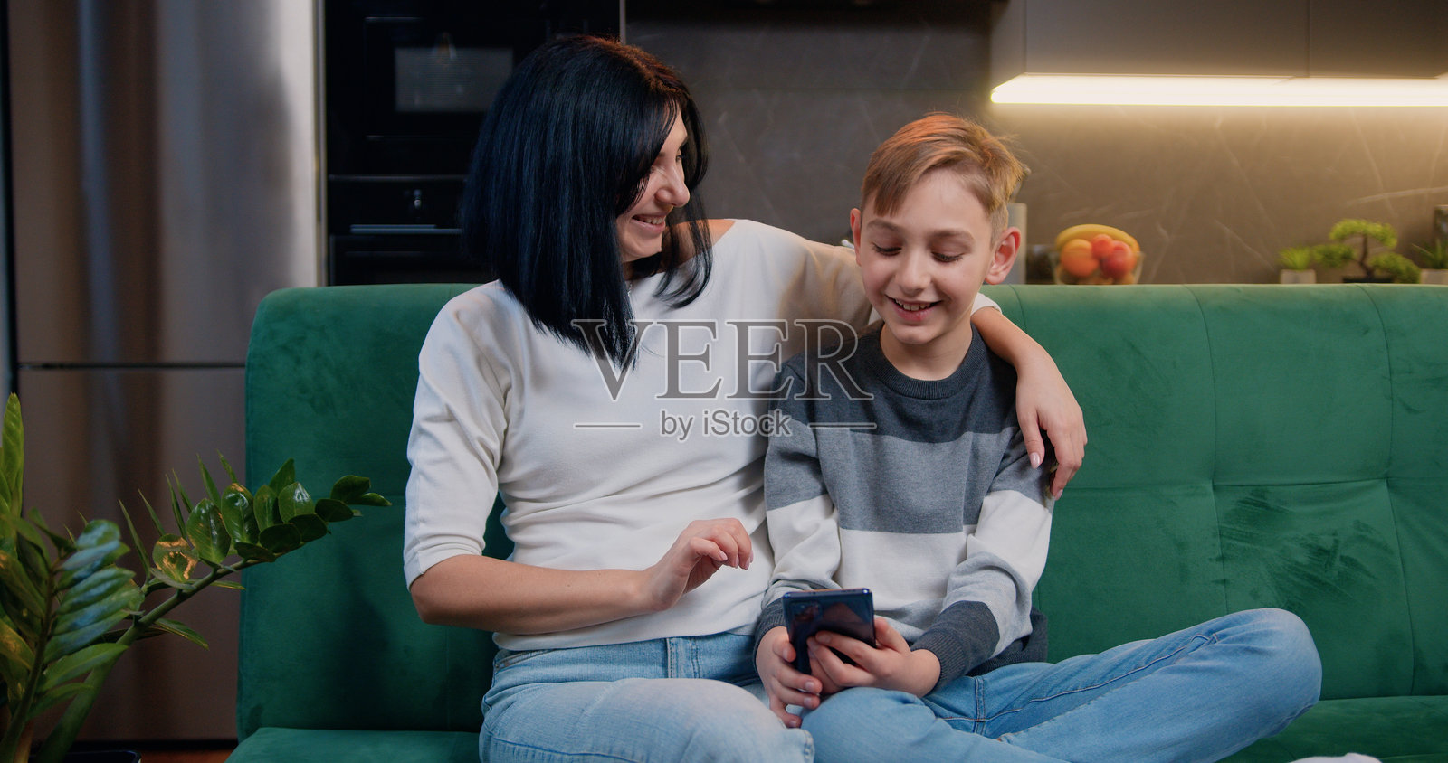 帅气的9岁白人男孩在沙发上与母亲一起玩智能手机，背景是厨房。设备，年轻一代互联网科技概念。照片摄影图片