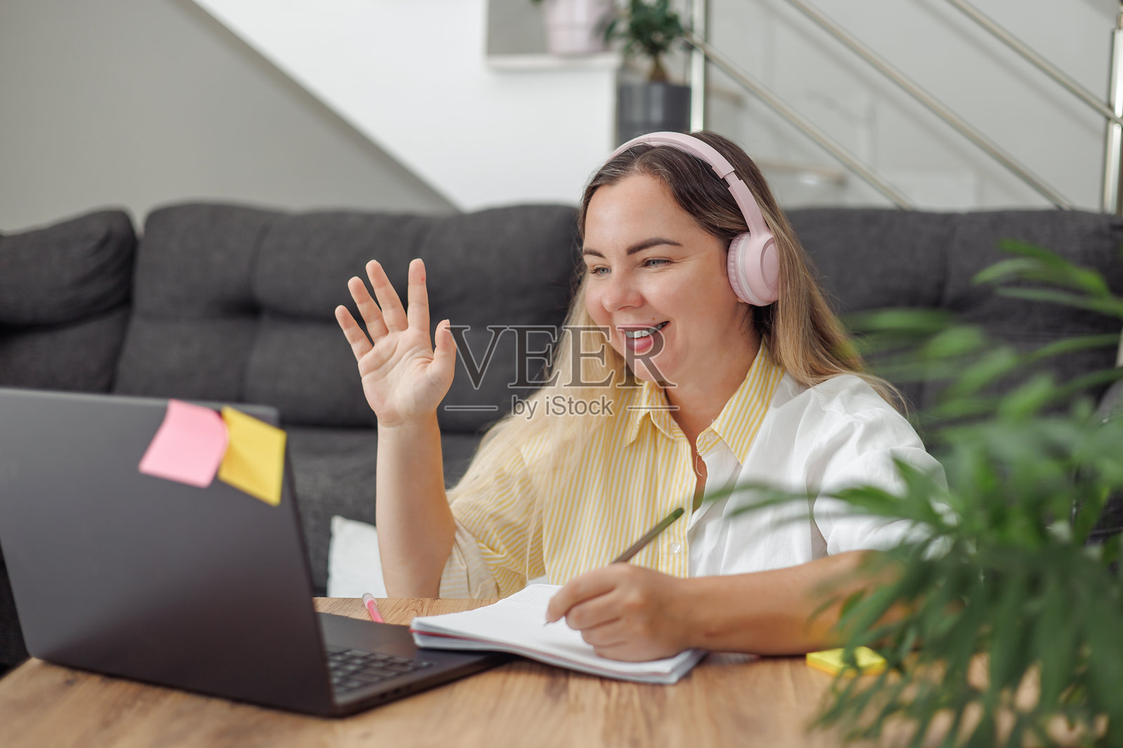 年轻的金发欧洲女性在家中与笔记本电脑和桌子上的笔记本进行视频通话时，戴着耳机并挥手。照片摄影图片