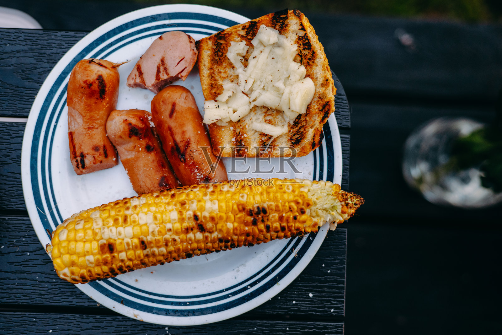 白色盘子上摆放着烤香肠、玉米和面包。这是一盘美味的烤食物，非常适合夏季烧烤。包括香肠、玉米棒和面包。照片摄影图片