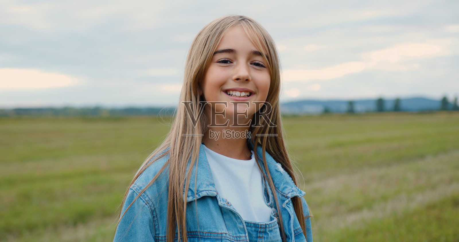 可爱的小金发女孩站在绿草地上，背景是美丽的日落。小女孩微笑着看向镜头，周围是迷人的自然风光。照片摄影图片