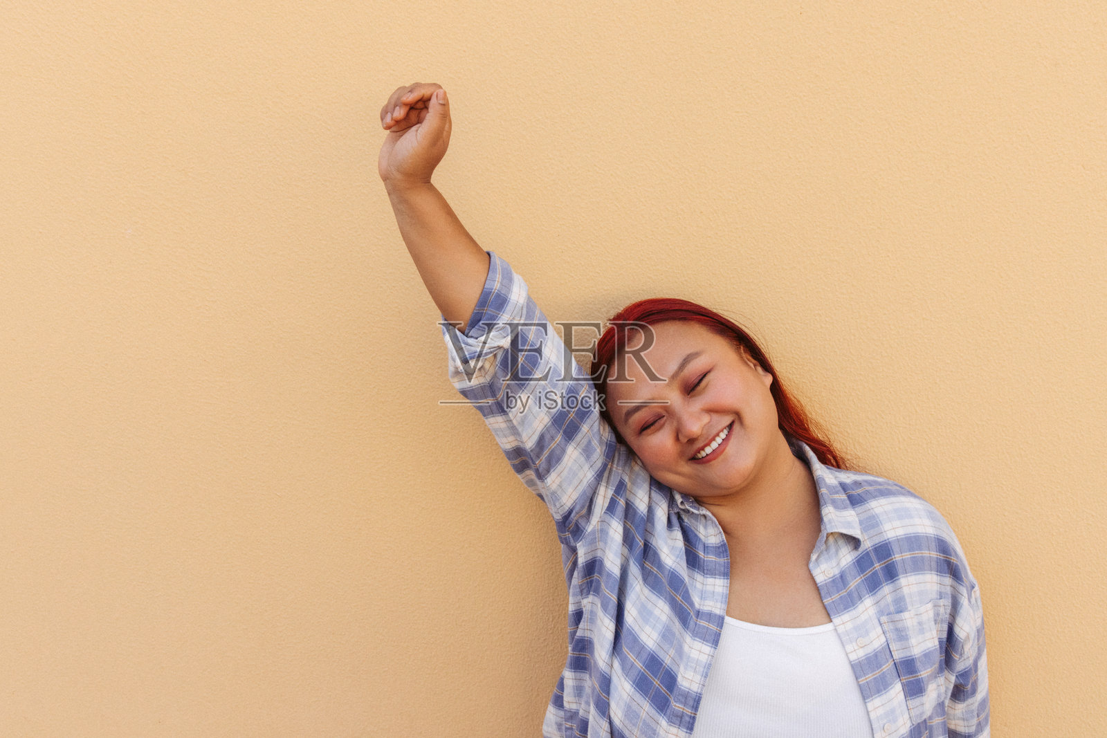 愉快的女性在浅橙色墙壁前微笑并举起手臂照片摄影图片