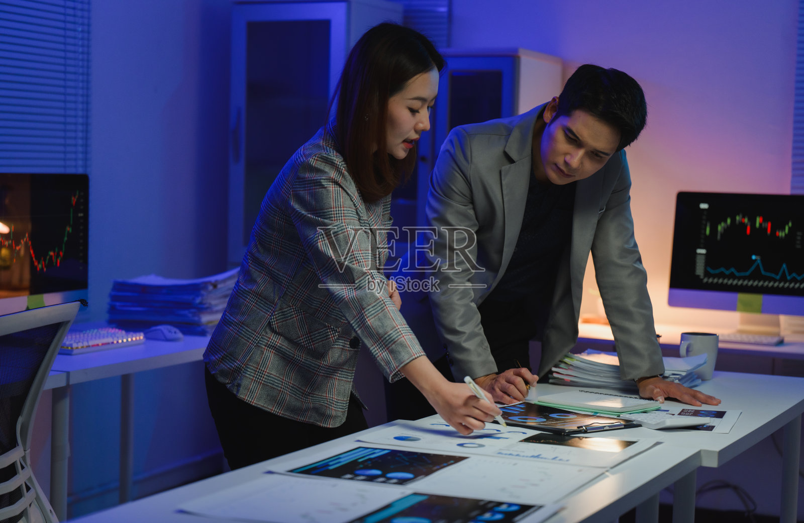 两位年轻人在现代办公室环境中合作进行财务分析。他们在柔和的灯光下查看纸上的图表和电脑屏幕上的数据。照片摄影图片