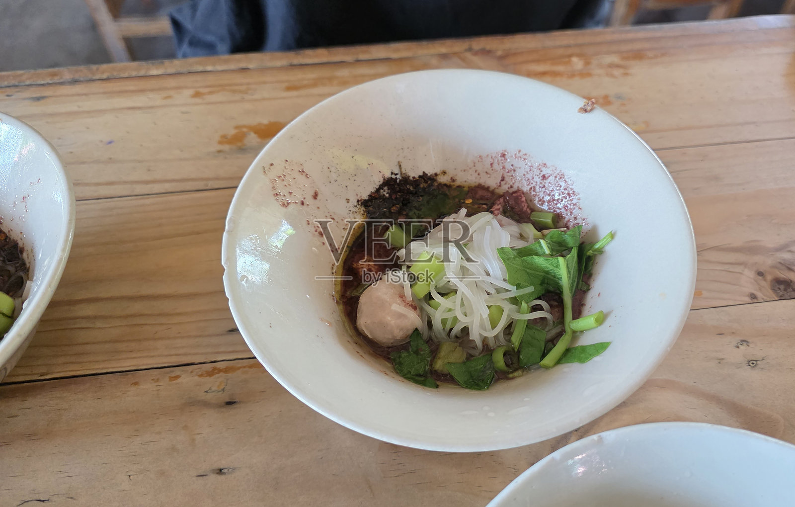 一碗传统泰国船面，配有米粉、猪肉片、青葱和浓郁的深色汤底，摆放在木桌上。照片摄影图片