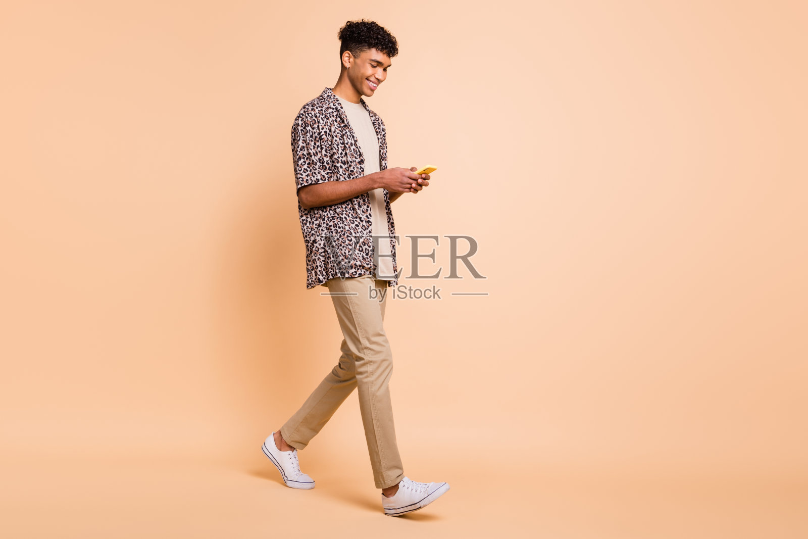 全身侧面照，棕发乐观男士，手持电话，穿着现代衬衫、长裤和运动鞋，背景为米色。照片摄影图片