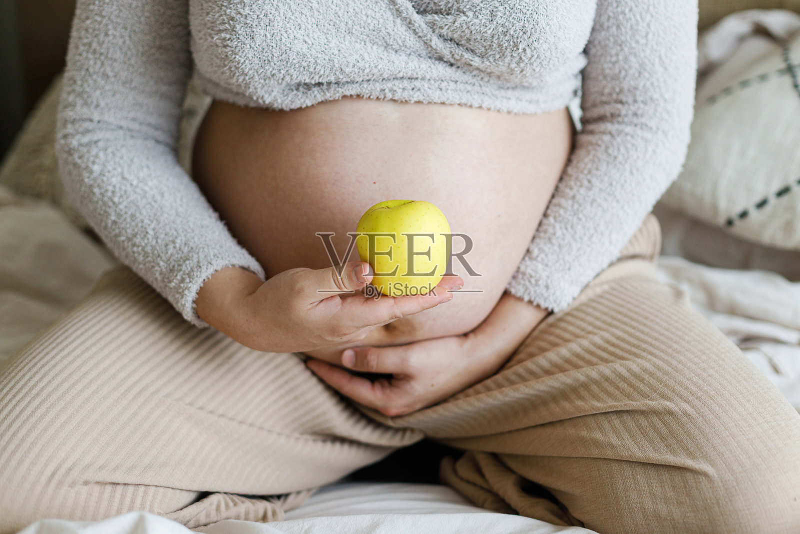 健康的怀孕与体重增加。孕妇坐在舒适的床上，抱着苹果，手放在肚子上。饮食与怀孕。照片摄影图片