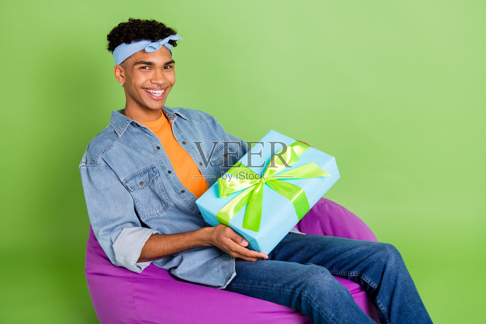 乐观友好的棕发男孩坐着，手里拿着礼物，穿着蓝色衬衫和牛仔裤，背景是绿色的。照片摄影图片