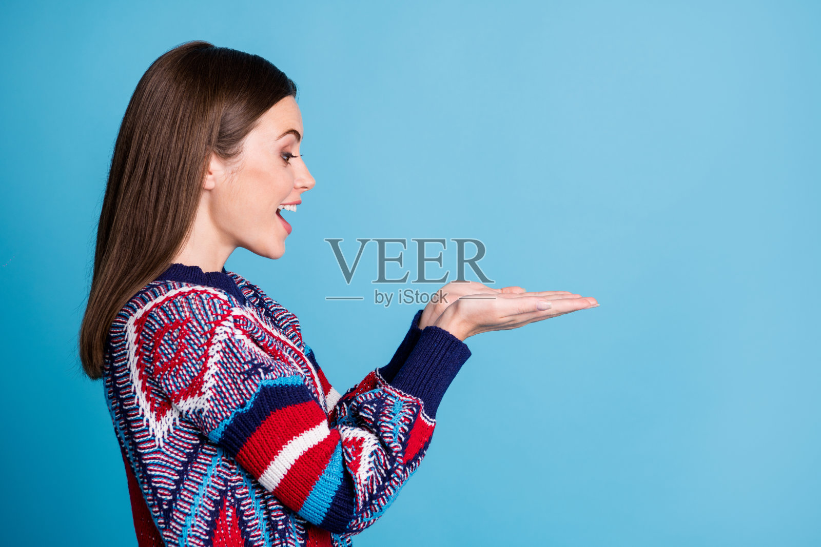 穿着毛衣的漂亮女孩侧面肖像照片，微笑着双手托着空白空间，背景为鲜艳的蓝色。照片摄影图片