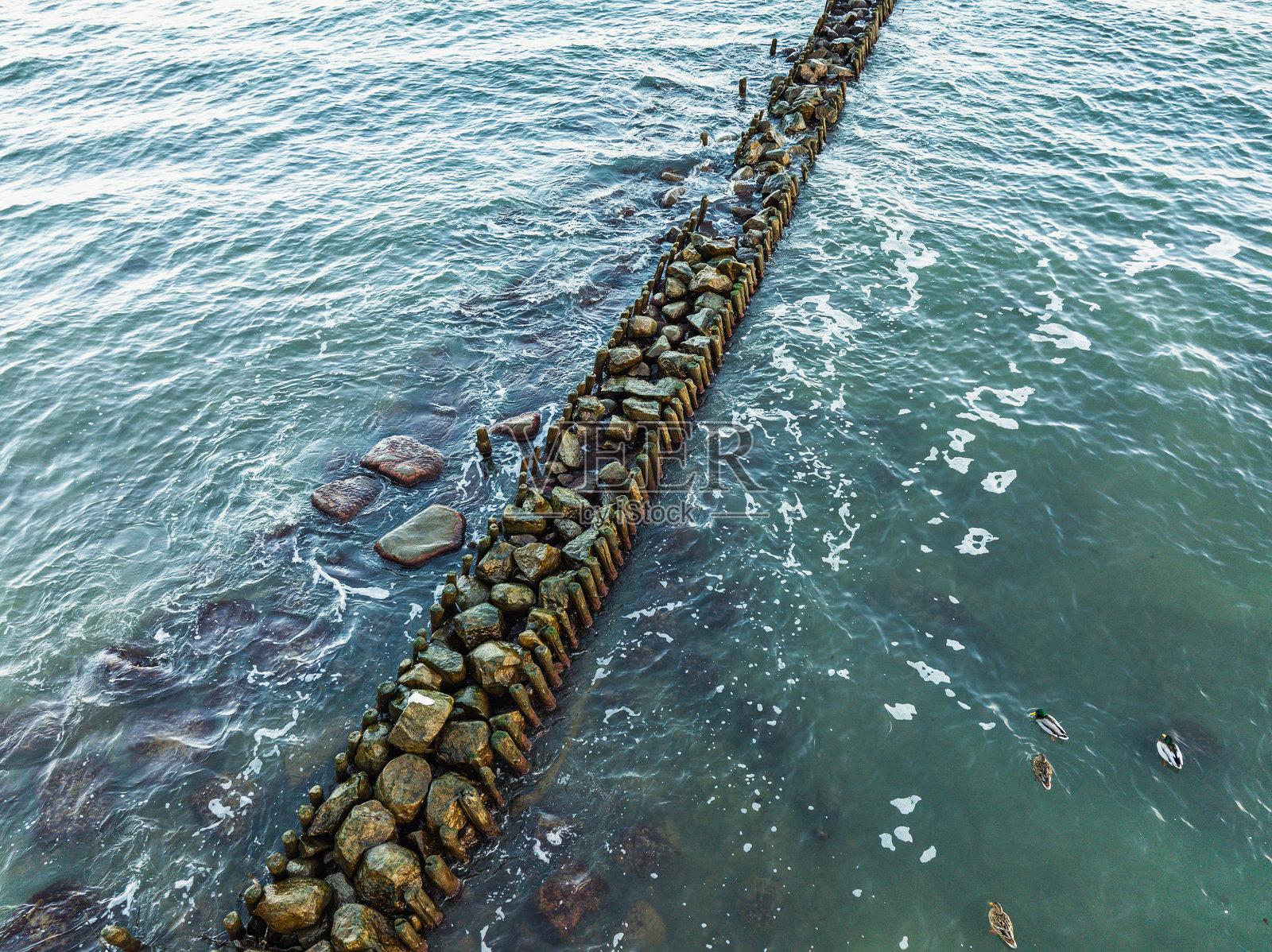 由大石块构成的防波堤延伸入清澈的蓝色海水中，为海岸提供保护。阳光在温柔的波浪上反射，营造出宁静的海岸氛围。照片摄影图片