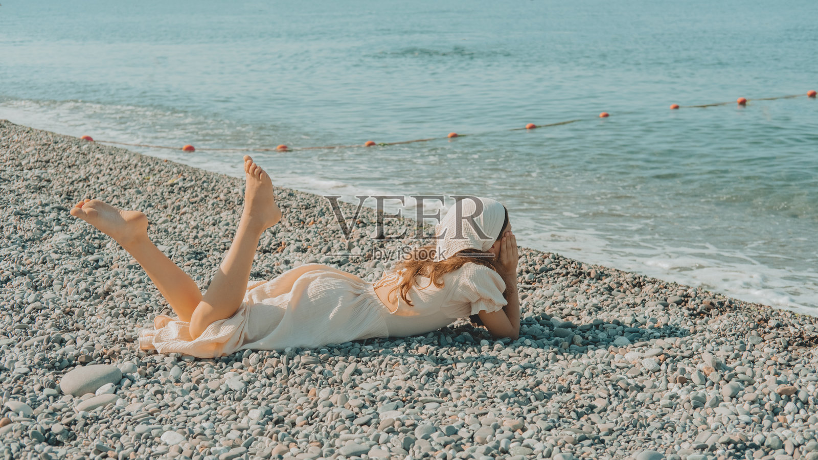 穿着轻盈裙子的女孩躺在卵石海滩上，双腿翘起，享受夏日的自由与宁静的海边氛围。照片摄影图片
