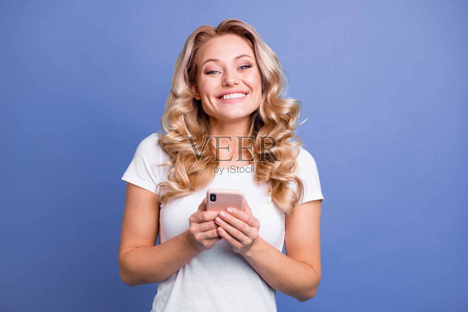 一位迷人、快乐、微笑的波浪卷发女孩在蓝色背景下使用社交媒体设备。照片摄影图片