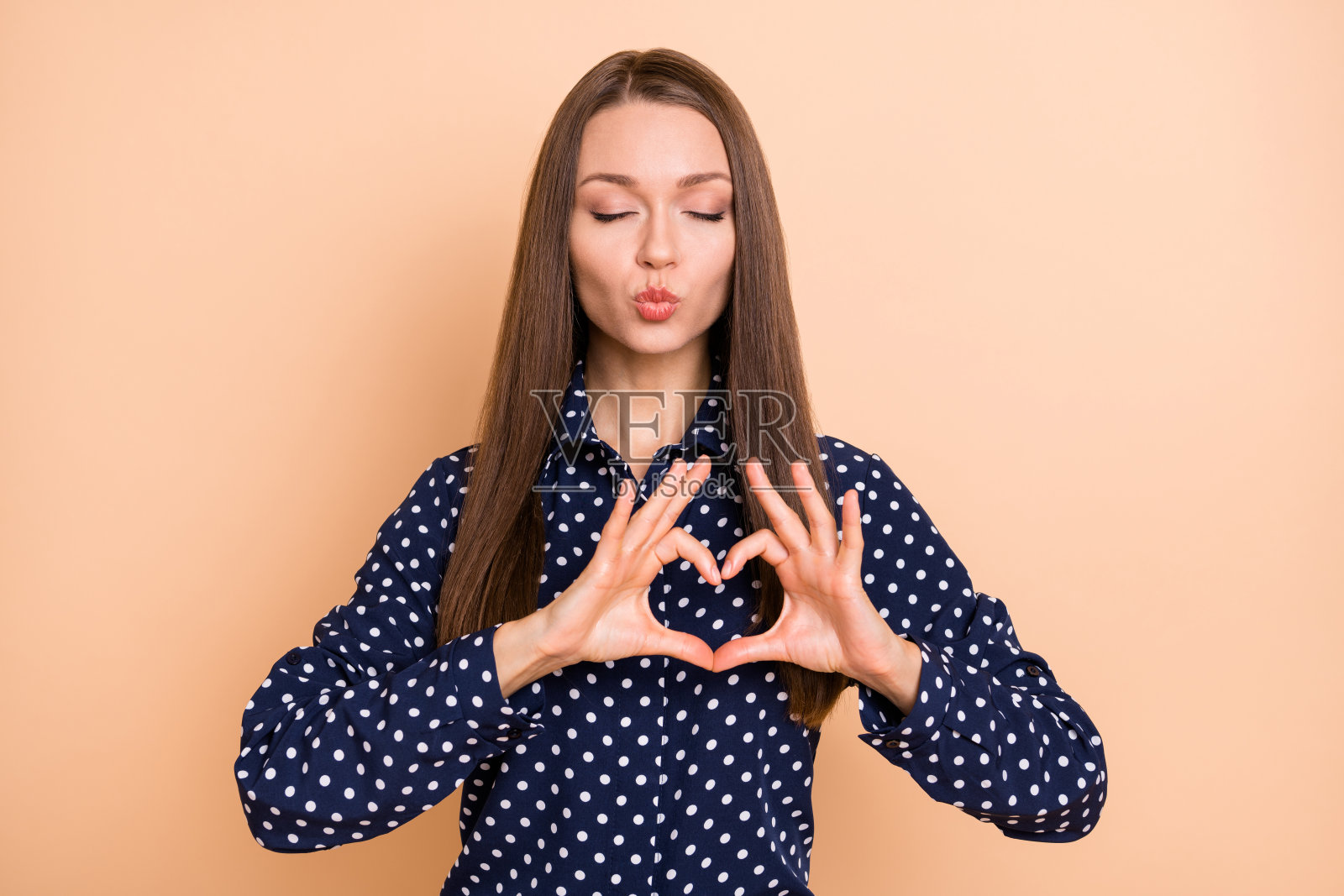 梦幻般的调皮年轻女孩穿着点点衬衫，闭着眼睛送出亲吻，背景为米色。照片摄影图片